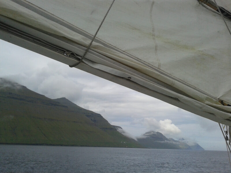 under a white bundle of sail, the triangular green cloud peaks of Kalsoy line up into the distance