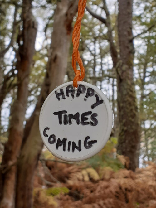 a round white disc hanging from a bright orange twine in the woods which reads 'happy times coming'