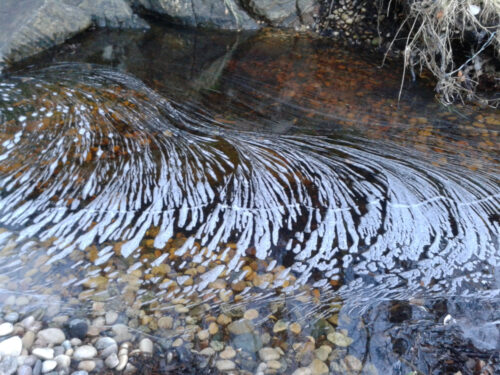 curled plumes of flat foam on brown water over pebbles